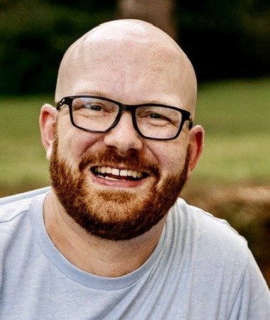 A smiling man with a bald head and glasses, wearing a light grey t-shirt, in an outdoor setting.